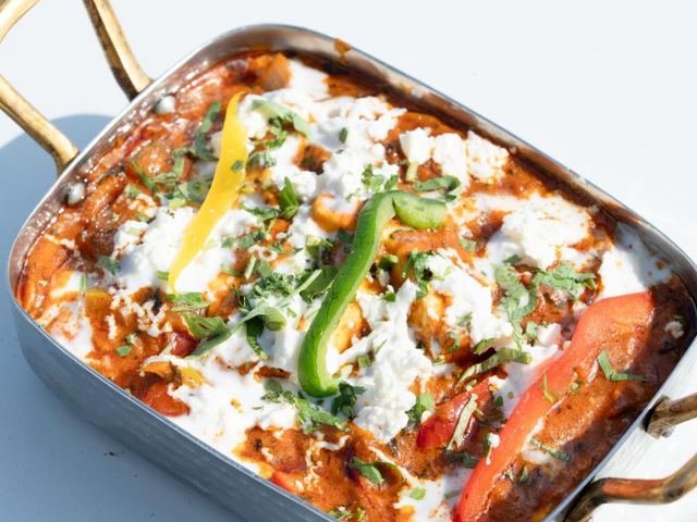 A rich tomato curry topped with cream, grated cheese and green chilli in a steel dish with brass handles.