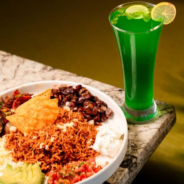 A rice bowl with beans, avocado, salsa and a tortilla chip, next to a bright green drink.