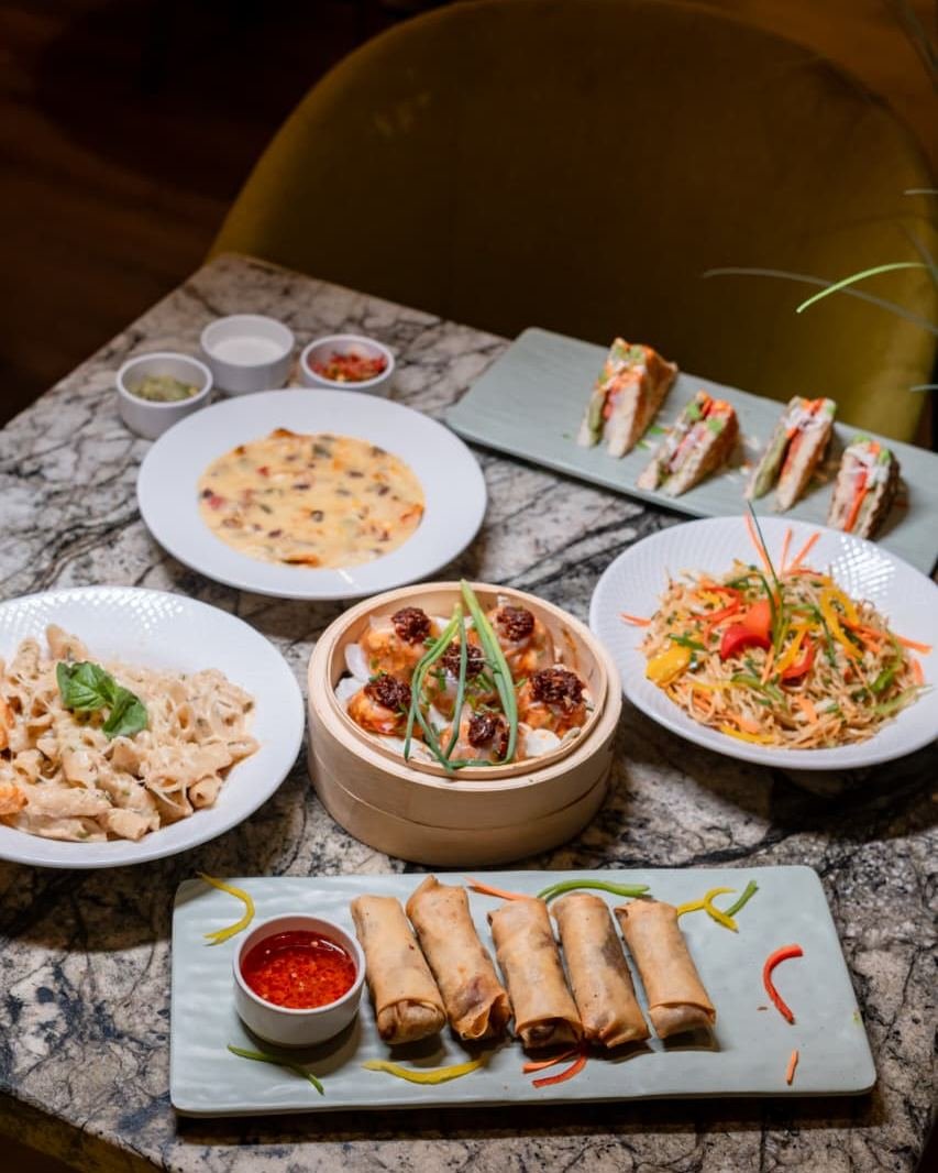 A marble table set with several dishes: spring rolls with a dip, stir-fried noodles, a bamboo steamer, toasted sandwiches and a bowl of soup.