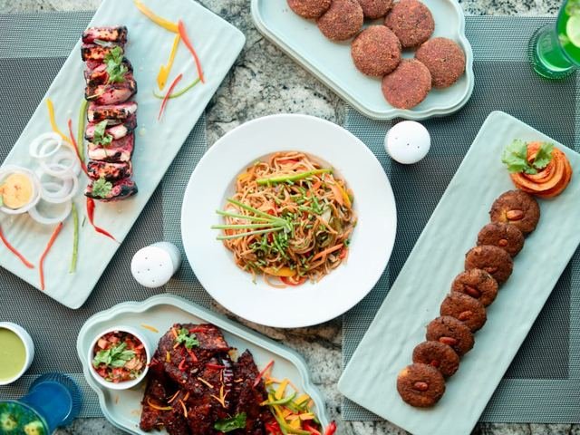 Overhead view of a table of plates: grilled skewers, a bowl of noodles, round kebabs, dips and drinks.